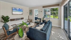 A modern living room with a sectional sofa, a coffee table, and a TV mounted on the wall. There is a large window providing a view of the outdoors.