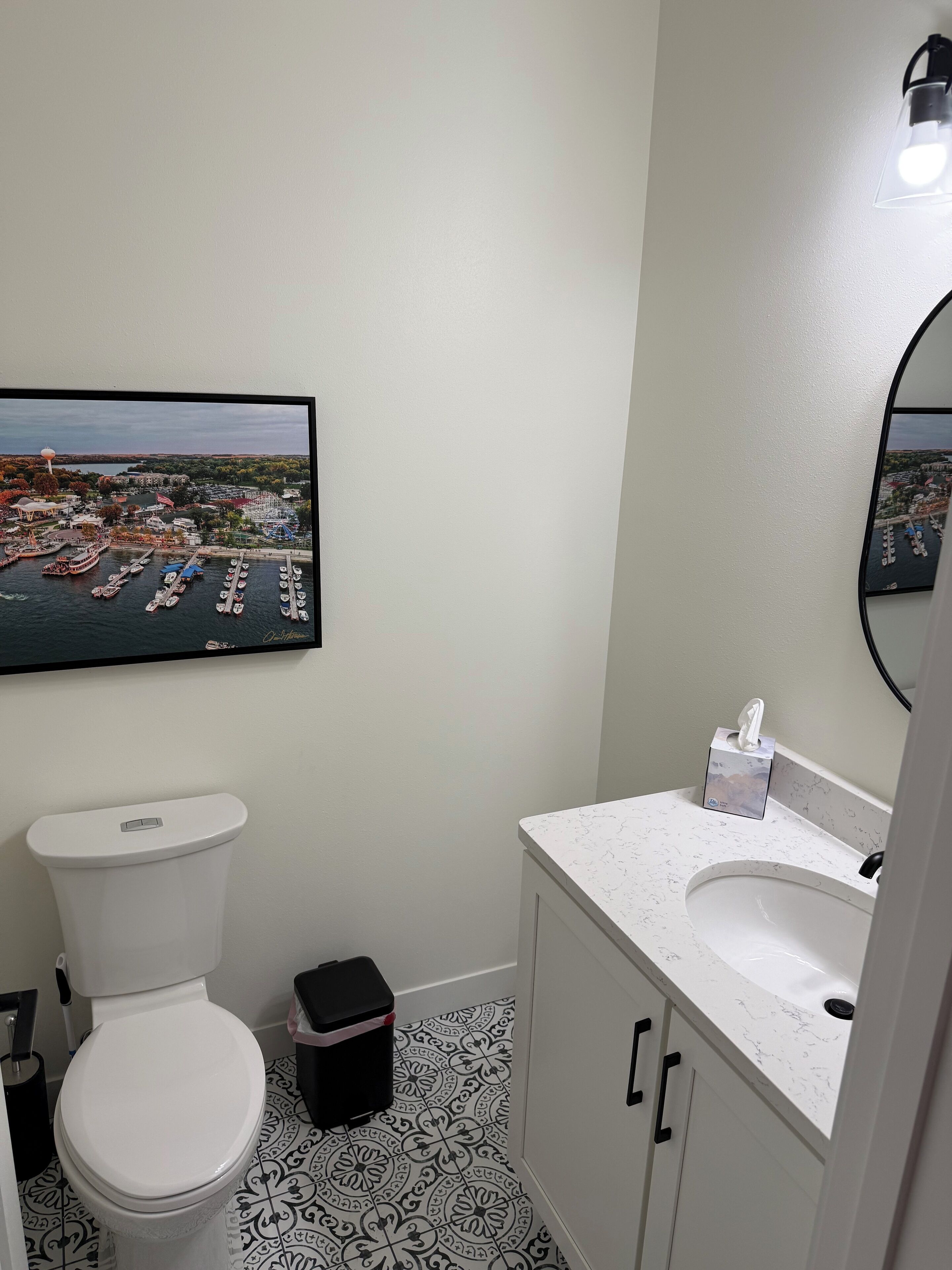 Un baño con inodoro, lavamanos y un espejo grande. Hay un cuadro en la pared con una imagen de un puerto deportivo.