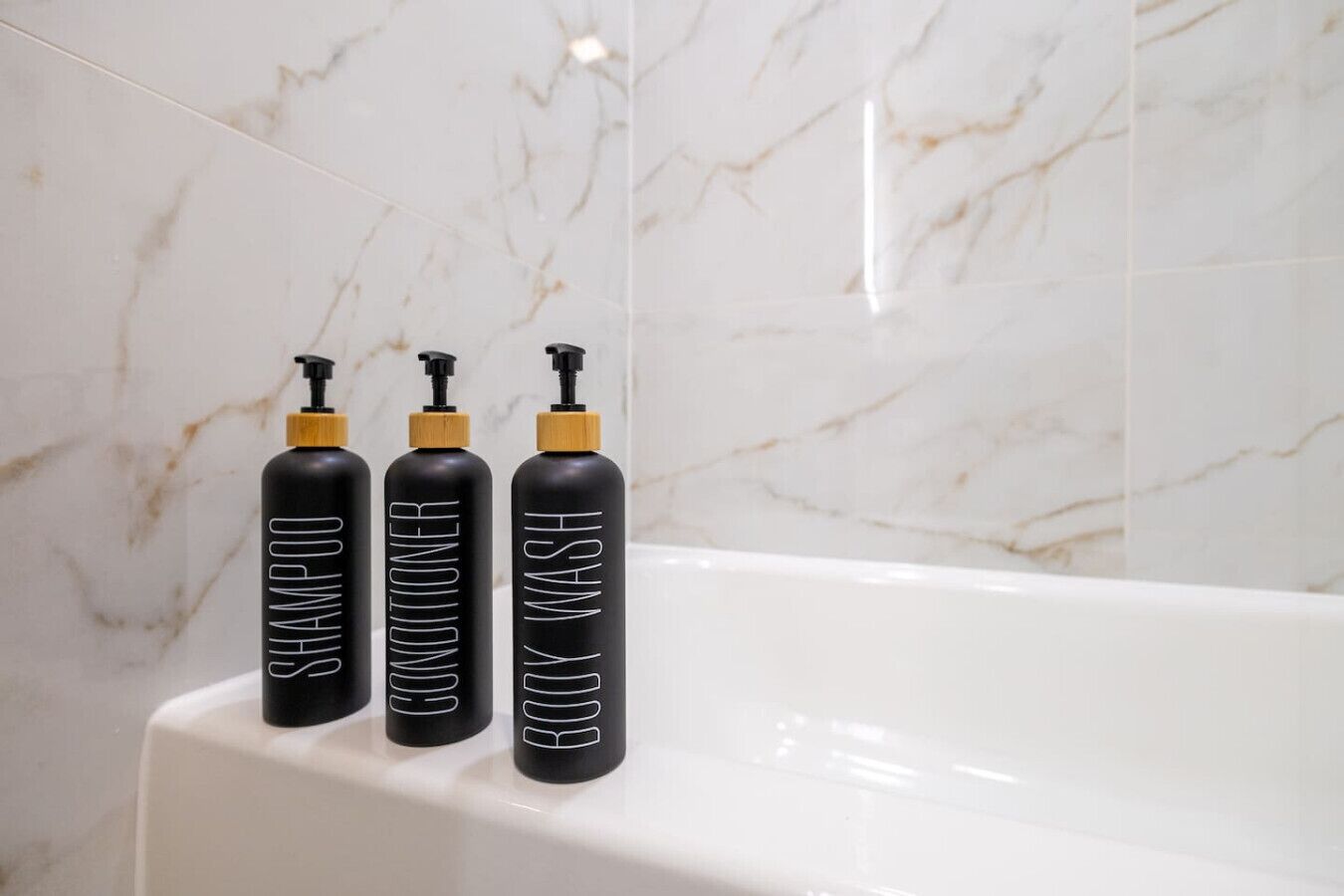 Trois flacons noirs équipés de pompes, portant les étiquettes « SHAMPOO », « APRÈS-SHAMPOING » et « GEL DOUCHE », sont posés sur une baignoire blanche dans une salle de bain aux carreaux de marbre.