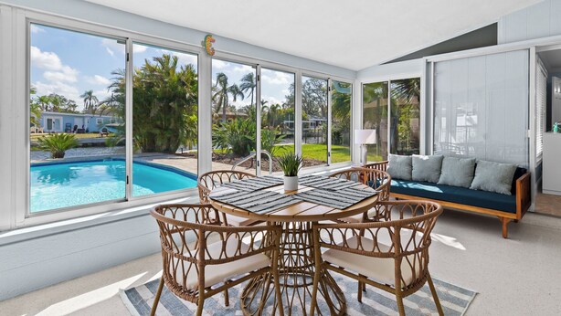 A spacious room with a large window overlooking a swimming pool and lush greenery. There is a round table with chairs and a sofa with cushions. - River House Retreat — Canal Views and Poolside Bliss! (Bradenton)
