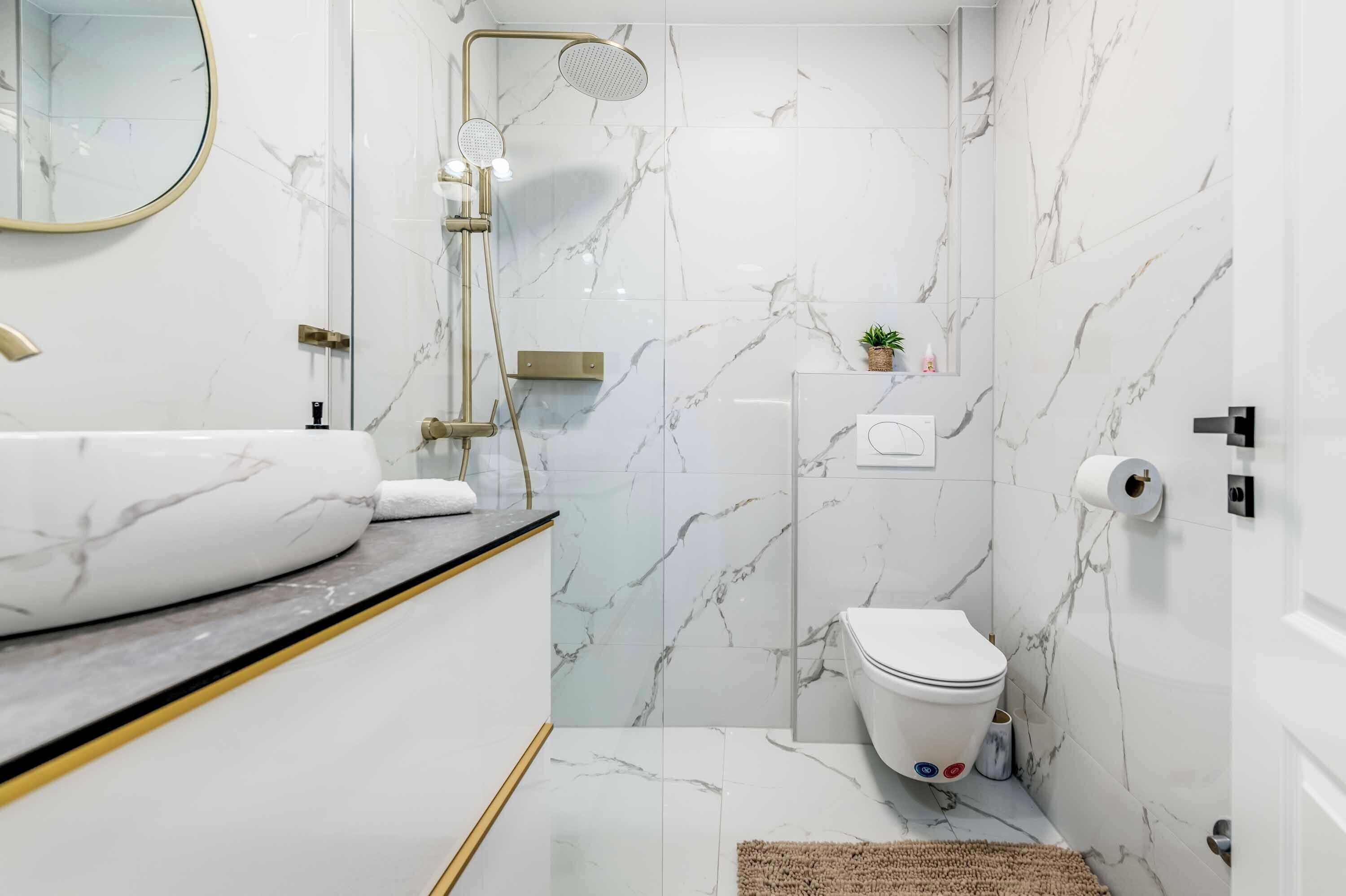 A modern bathroom with a marble floor, a large white sink, a toilet, and a shower with a gold showerhead.