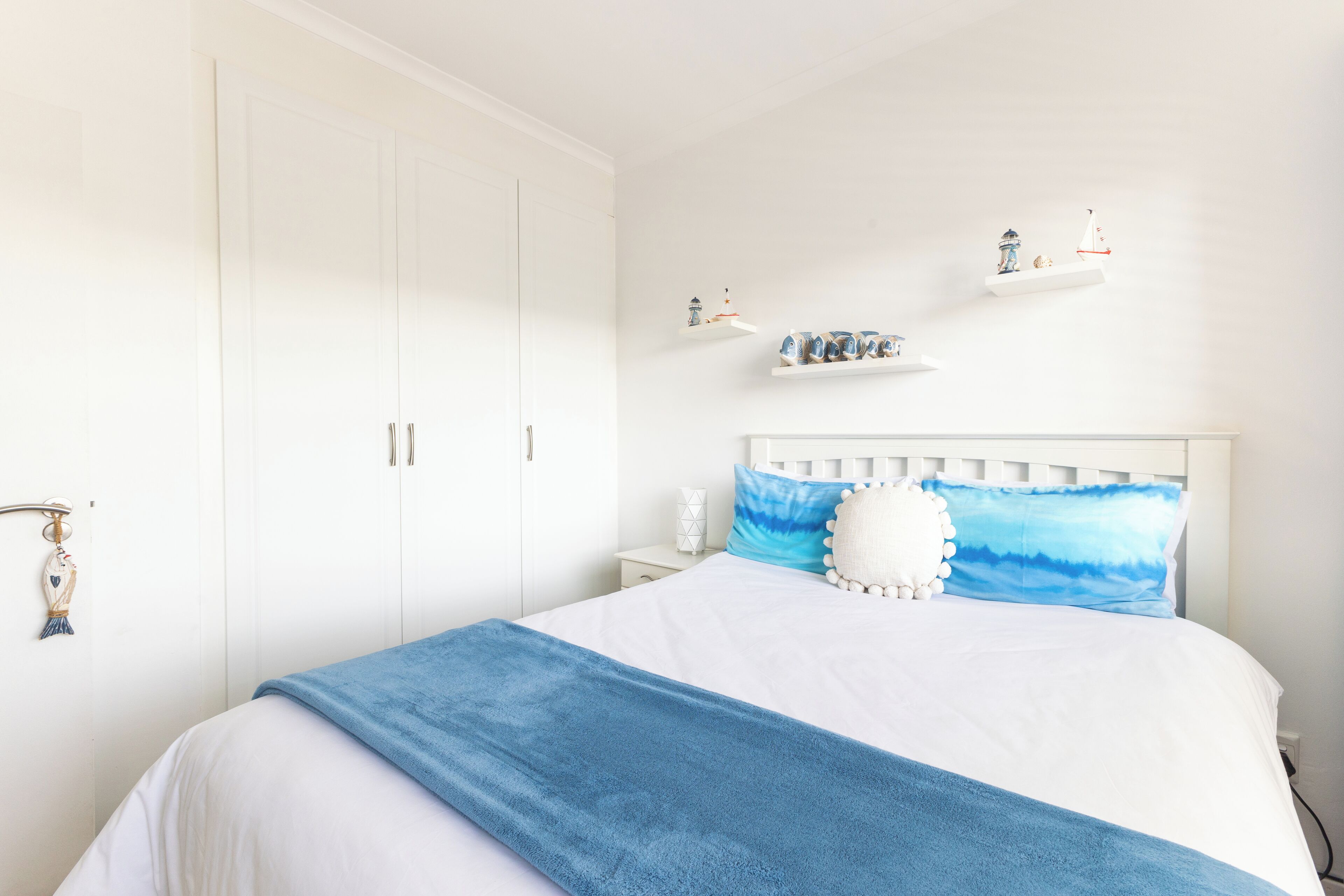 A bedroom with a white wardrobe, a white headboard, and a bed with a blue blanket. There are shelves with decorative items on the wall.