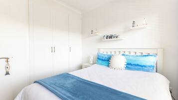A bedroom with a white wardrobe, a white headboard, and a bed with a blue blanket. There are shelves with decorative items on the wall.