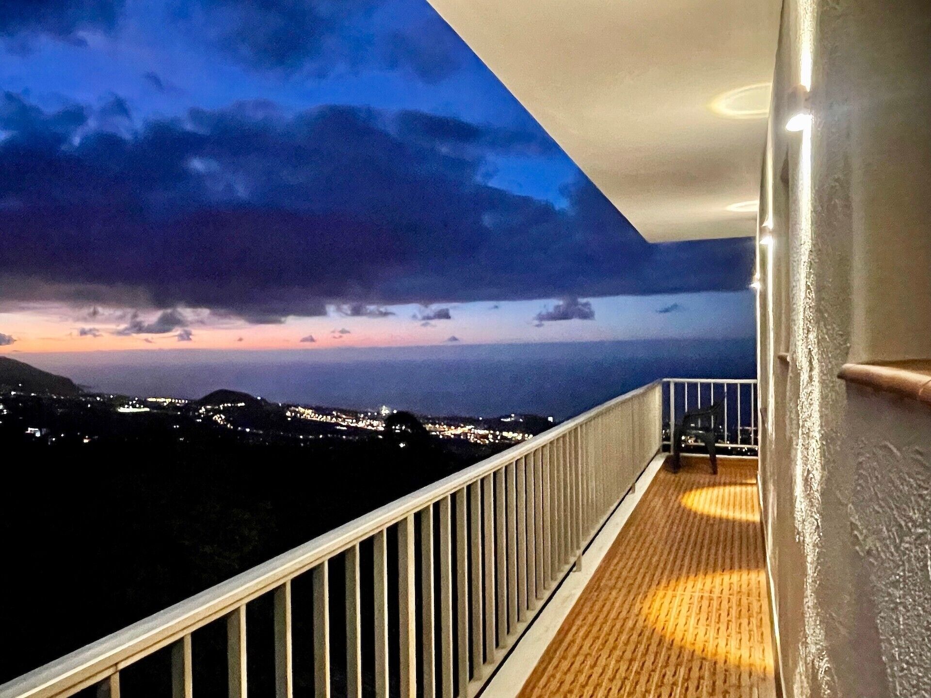 Un balcón con barandilla, piso de madera y vista a un paisaje urbano al anochecer.