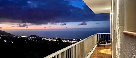 A balcony with a railing, a wooden floor, and a view of a cityscape at dusk.