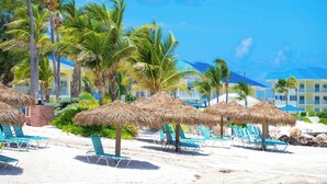 Una playa con sombrillas de paja, sillas de descanso, palmeras y un hotel al fondo.