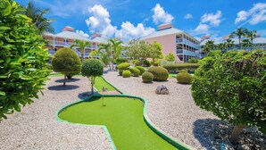 Un campo de golf con putting green, rodeado de arbustos y árboles bien cuidados, y un hotel al fondo.