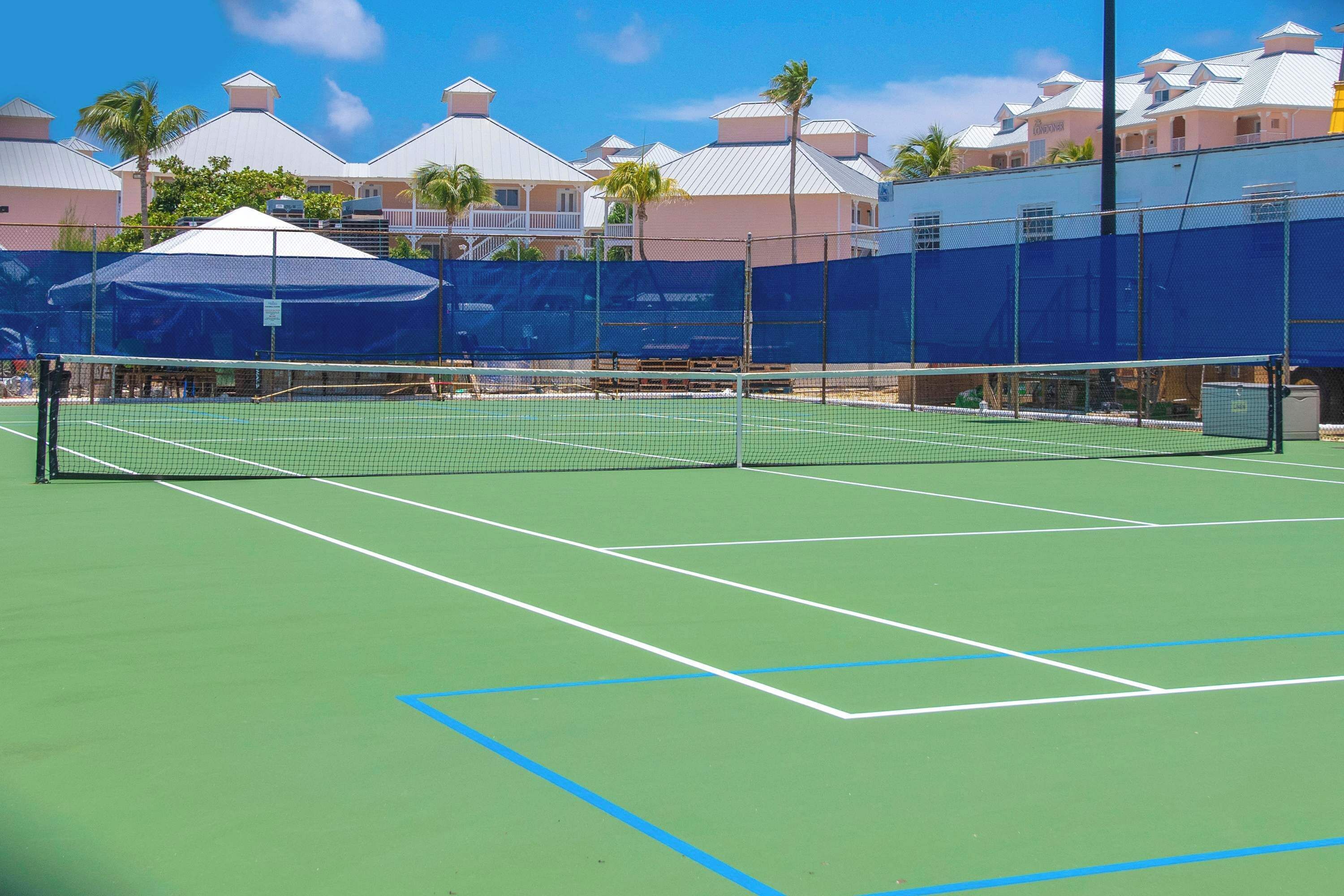 Ein Tennisplatz mit Netz, umgeben von einem Zaun, und Gebäuden im Hintergrund.
