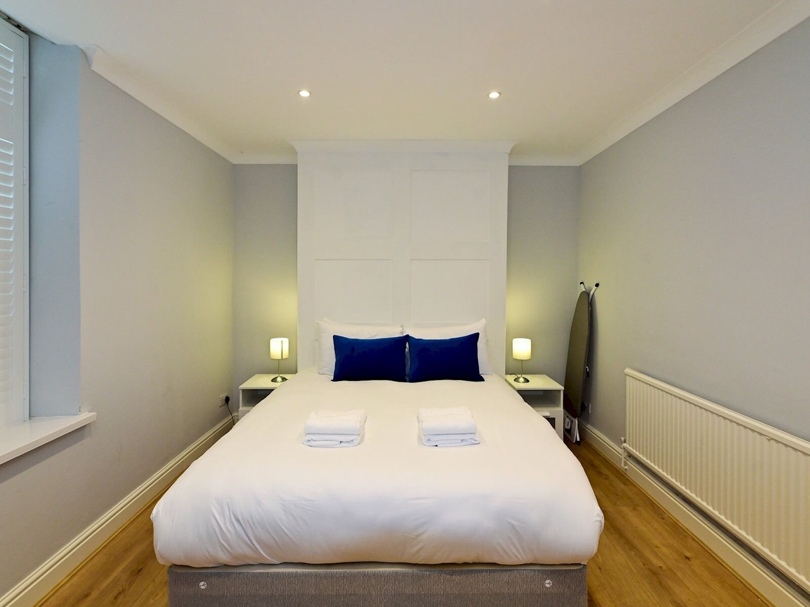 A neatly made bed with white linens and two folded towels, a bedside table with a lamp, a radiator, and a window with blinds.