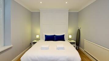 A neatly made bed with white linens and two folded towels, a bedside table with a lamp, a radiator, and a window with blinds.