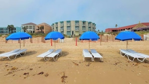 A beach with lounge chairs and blue umbrellas, a building with a round shape, and a red building with a thatched roof in the background. - Block to Beach - Pool - Up to 6 - Families (South Padre Island)