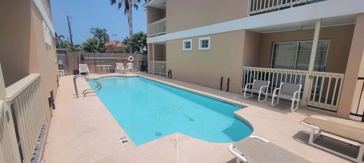 A swimming pool with lounge chairs and a fenced area, surrounded by a building with balconies.