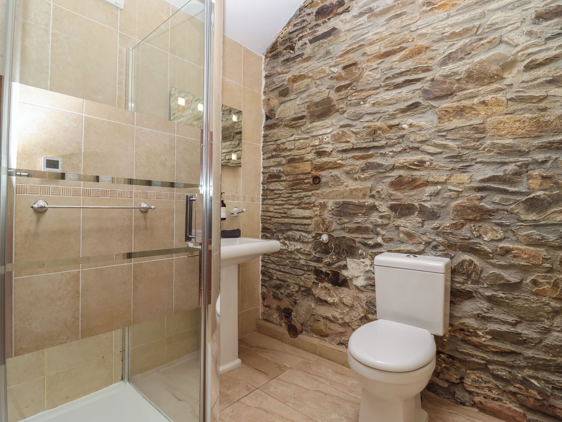 Un baño con ducha envuelta en vidrio, un lavamanos y un inodoro frente a una pared de piedra.