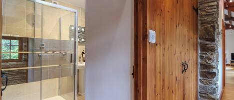 A modern bathroom with a glass shower enclosure, a wooden door with a stone accent, and a visible light switch.
