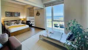 A hotel room with a bed, a sofa, a chair, a plant, and a large window with a view of the sea. - MactanNewtown Staycation Near The Beach  (Lapu-Lapu)
