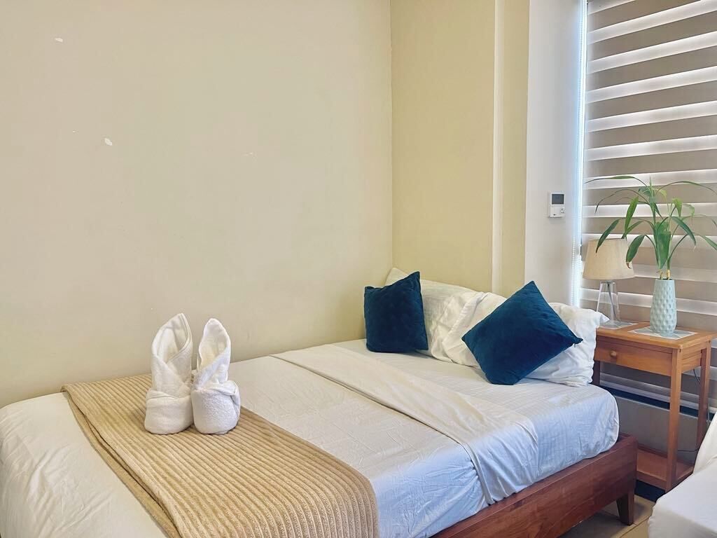Un letto rifatto con una coperta beige, due cuscini e un paio di pantofole bianche sul lato.