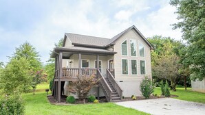 A two-story house with a front porch, a staircase leading to the second floor, and a small front yard with shrubs and a tree.