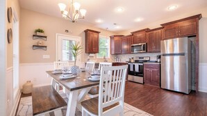 A modern kitchen with wooden cabinets, stainless steel appliances, and a dining area with white chairs and a wooden table.
