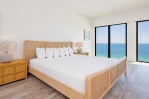 A modern bedroom with a large bed, wooden headboard, bedside tables, and a view of the ocean.
