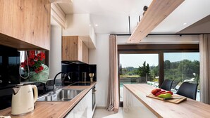 A modern kitchen with wooden cabinets, a stainless steel sink, and a wooden countertop. There is a large window with a view of trees and a balcony with outdoor seating.