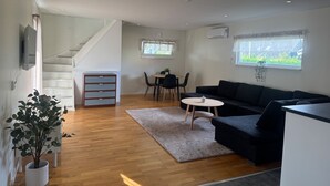 A modern living room with a black sofa, a coffee table, and a dining area with a table and chairs. There is a staircase leading to another level, and a plant in a white pot is visible. - Modern 1-bedroom Apartment for 6 Near Kristianstad (Fjalkinge)