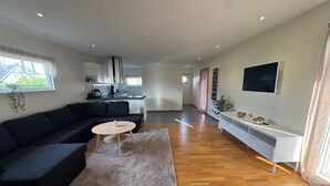 A modern living room with a dark-colored sectional sofa, a round coffee table, and a white console table with decorative items. A kitchen area is visible in the background with white cabinets and a large window. - Modern 1-bedroom Apartment for 6 Near Kristianstad (Fjalkinge)