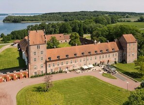 A large, historic building with multiple wings, surrounded by well-maintained lawns and a body of water in the background.