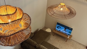 A modern living room with a brown sofa, two wicker lamps, a small table with a TV, and a patterned stool.
