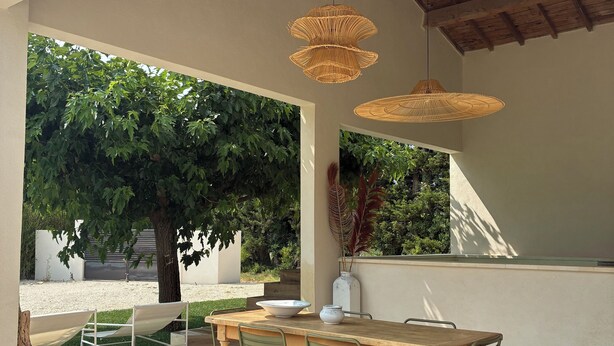 An outdoor patio with a wooden table and chairs, surrounded by a garden with trees and a view of the landscape.