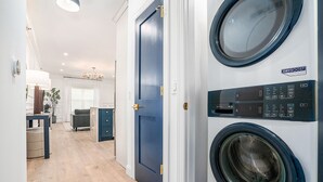 A laundry room with two front-loading washing machines, a blue door, and a hallway leading to a living area with a chandelier.