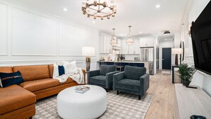 A modern living room with a brown sofa, grey armchairs, and a white ottoman. There is a kitchen in the background with white cabinets and a chandelier.