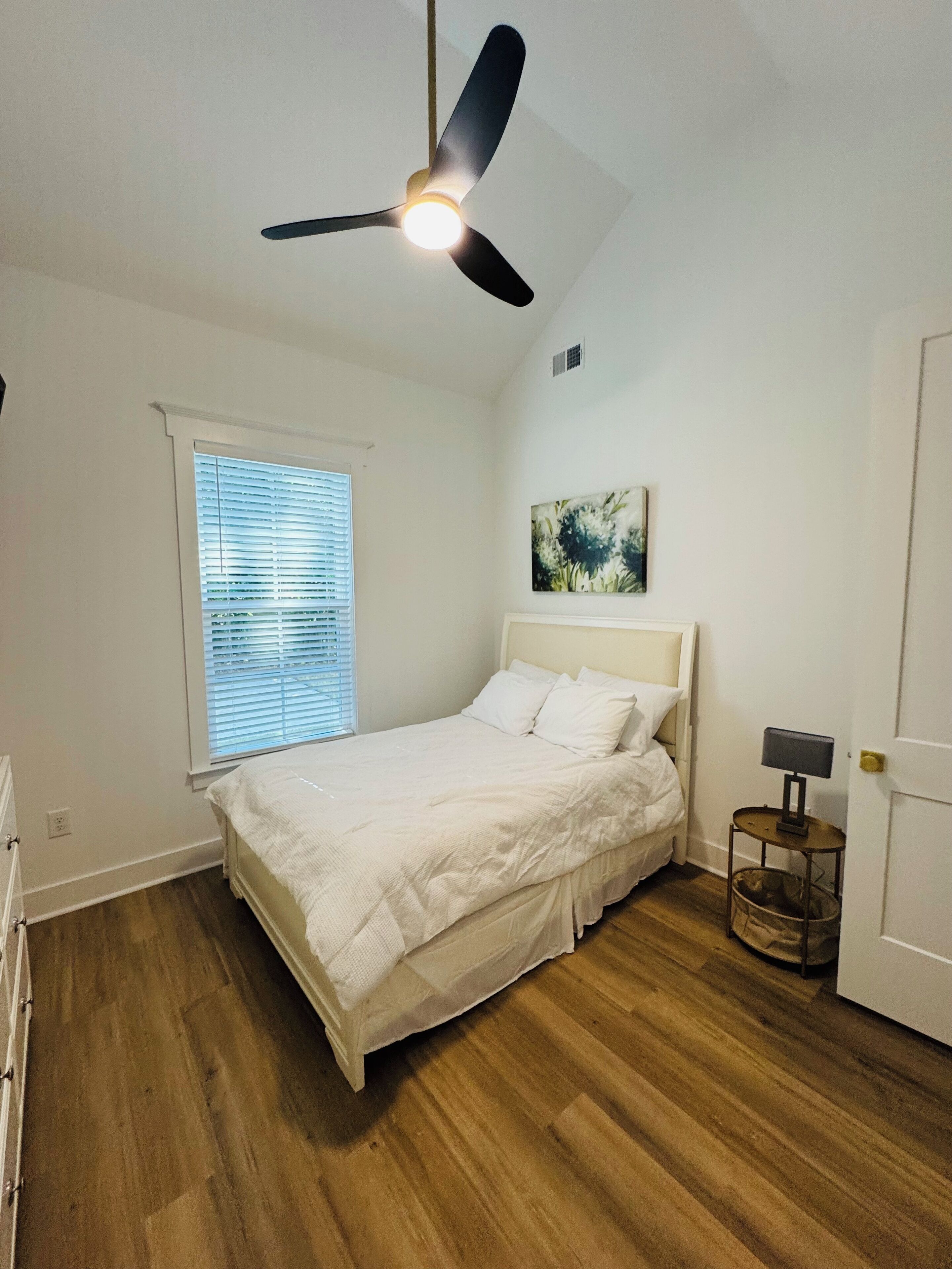 Ein Schlafzimmer mit einem Bett, einem Deckenventilator, einem Fenster mit Jalousien, einem Bild an der Wand und einem kleinen Tisch mit Lampe.