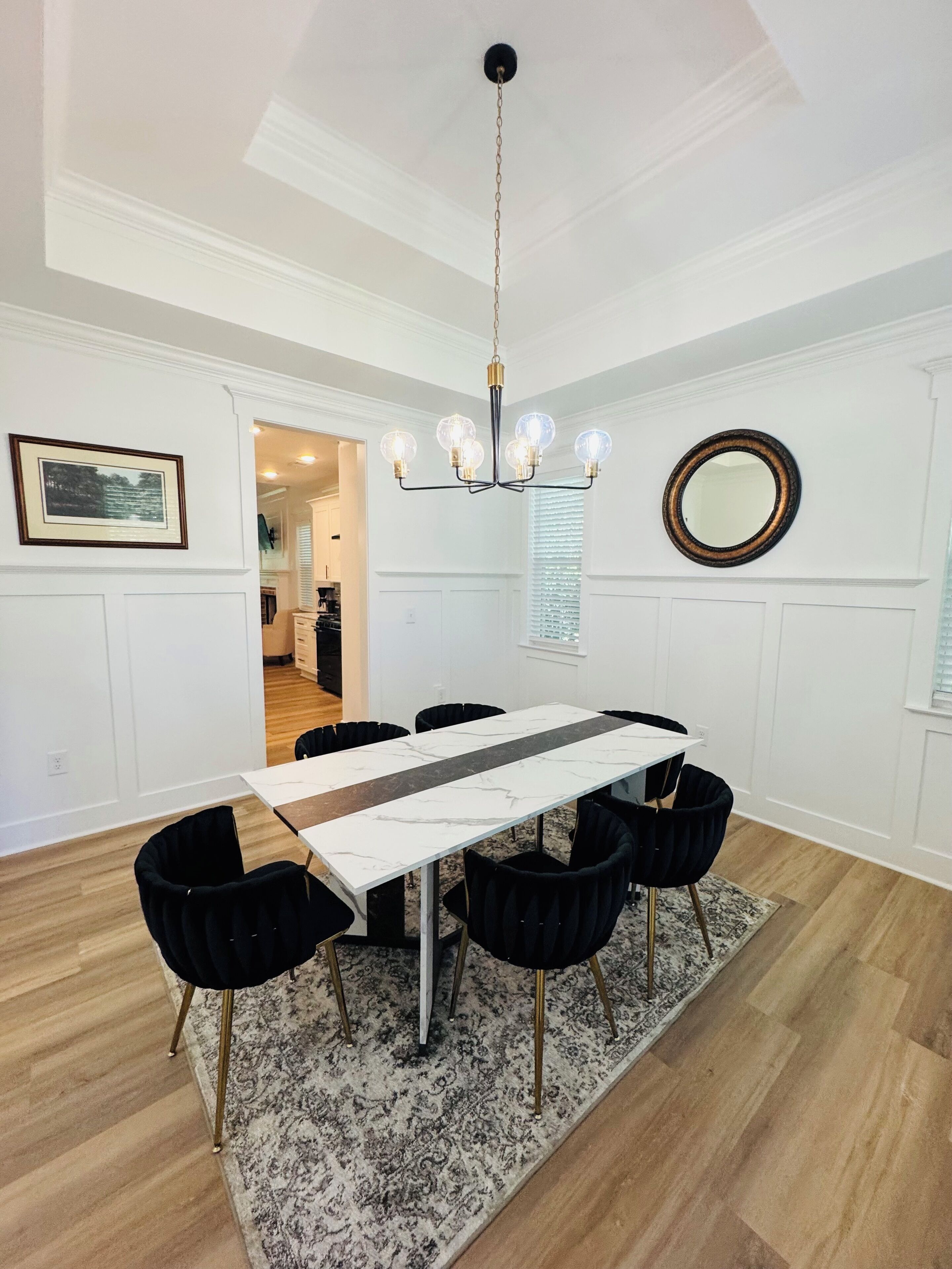 A dining area with a marble-top table, black chairs, a patterned rug, a chandelier, and a mirror on the wall.