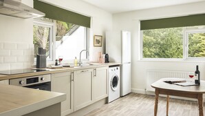 A modern kitchen with a dishwasher, oven, and washing machine. There is a window with a view of greenery outside. - Tethers End (Windermere)