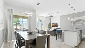 Une cuisine moderne avec des meubles blancs, un coin repas doté d’une grande table et de chaises, et un salon avec un canapé et un téléviseur.