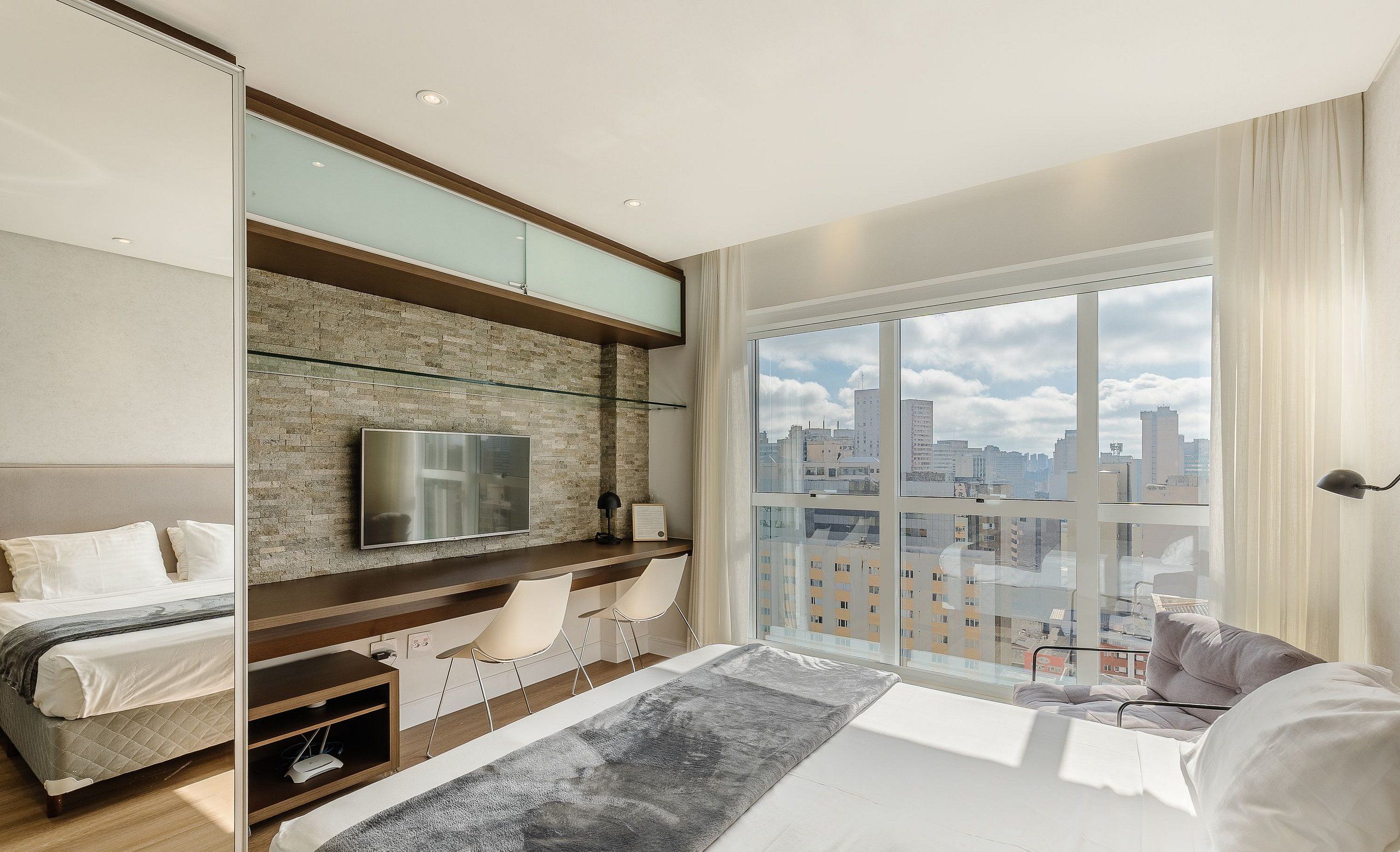 A modern bedroom with a bed, a TV, a desk, and a large window with a city view.