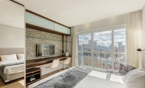 Quarto moderno com cama, TV, escrivaninha e uma grande janela com vista para a cidade.