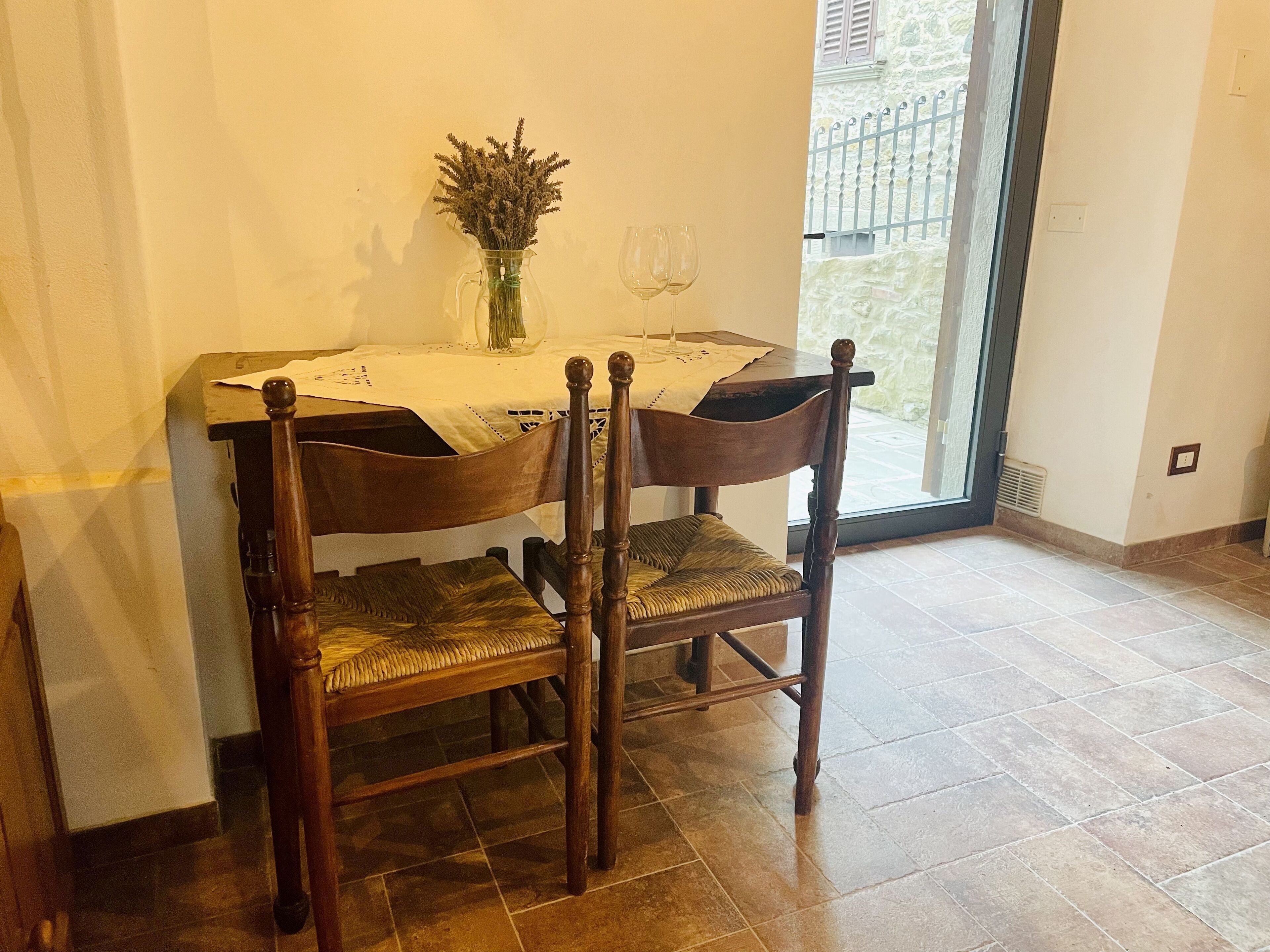 A dining area with a table set for two, featuring a vase with dried flowers, two wine glasses, and a wooden chair with a woven seat.