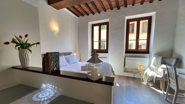 A bedroom with a bed, bedside table, lamp, chair, and a view of the outside through windows.