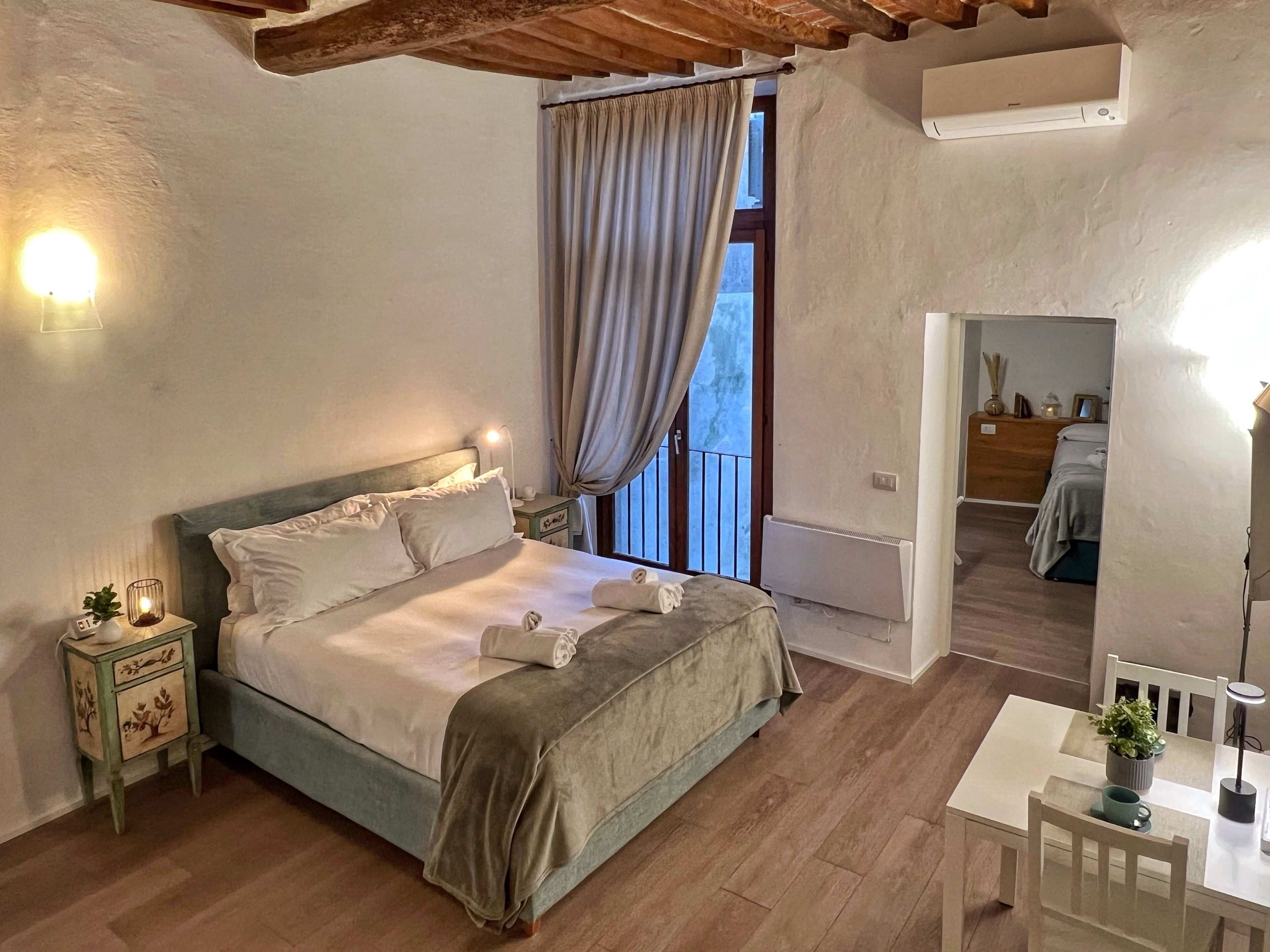 Un dormitorio con cama, mesitas de noche, una ventana con cortinas, un radiador, una pequeña mesa con una planta y una silla.