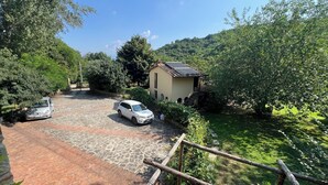 Une allée pavée où sont garées deux voitures, un petit bâtiment avec un toit en tuiles et une végétation luxuriante qui entoure la propriété.