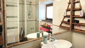 Une salle de bain avec une douche à parois de verre, un lavabo blanc, un miroir et des étagères en bois où sont rangés les articles de toilette.