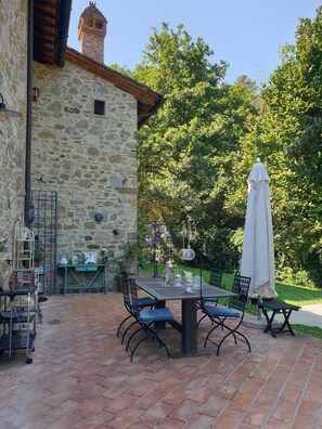 A stone building with a rustic exterior, a patio area with a table and chairs, and a garden with trees and plants.