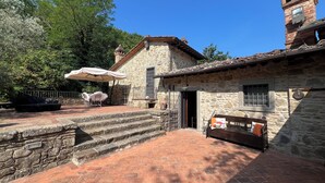 Un edificio de piedra con techo de tejas, un pequeño balcón y un patio empedrado con mobiliario exterior.