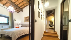 Un dormitorio con cama, mesita de noche, lámpara y ventana. Se ve un pasillo que lleva a un comedor.
