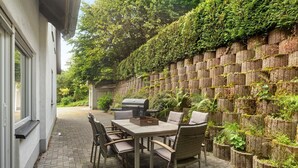 An outdoor patio with a table and chairs, a grill, and a wall with vertical planters. - Holiday Home Talblick / 160 m² / Up to 8 Guests / Garden / 2 Bathrooms / Sauerland (Schmallenberg)