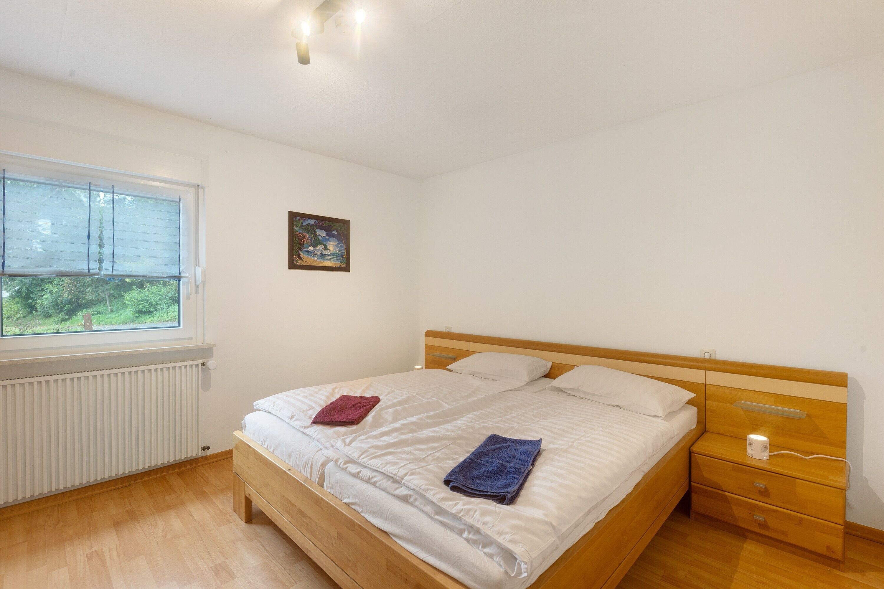 Une chambre à coucher avec un lit, une table de chevet, une fenêtre, un radiateur et un tableau au mur.