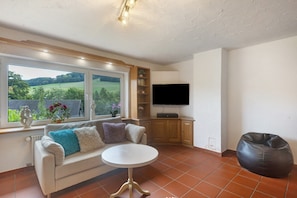 Un soggiorno con divano, un tavolino rotondo, un pouf, una TV e una finestra con vista sul verde.