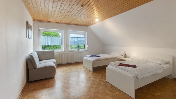 A bedroom with a wooden ceiling, two beds, a sofa, and two windows with curtains. - Holiday Home Talblick / 160 m² / Up to 8 Guests / Garden / 2 Bathrooms / Sauerland (Schmallenberg)
