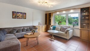 A living room with a sofa, a round table, a white sofa, a bookshelf, and a window with a view of greenery. - Holiday Home Talblick / 160 m² / Up to 8 Guests / Garden / 2 Bathrooms / Sauerland (Schmallenberg)